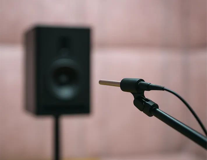 A close-up of a microphone stand with a cable, in front of a black studio monitor speaker against a light pink wall.